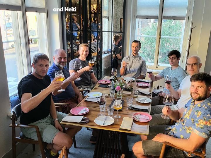 Seven men smiling and raising glasses while posing for a group photo during a team dinner at a restaurant, with a set dining table.