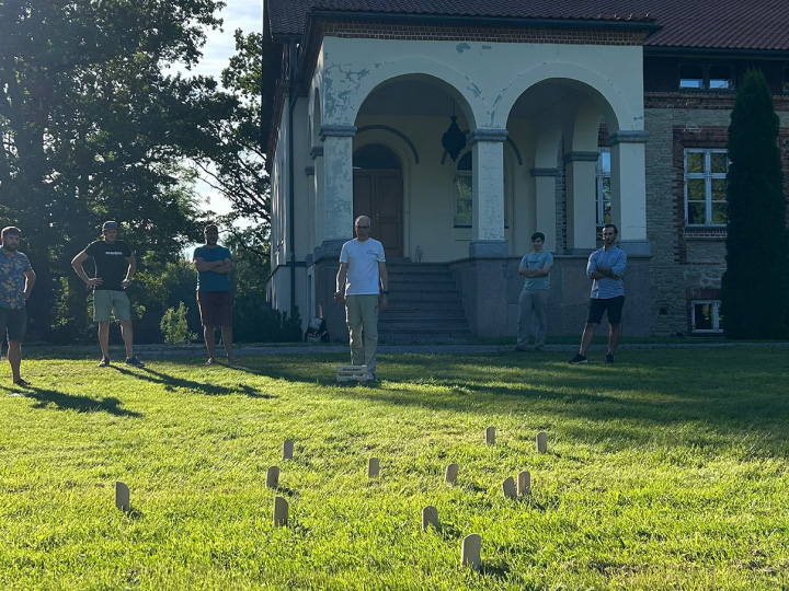 Six men are playing molkky in an outdoor setting, grass is green and the sun is shining.