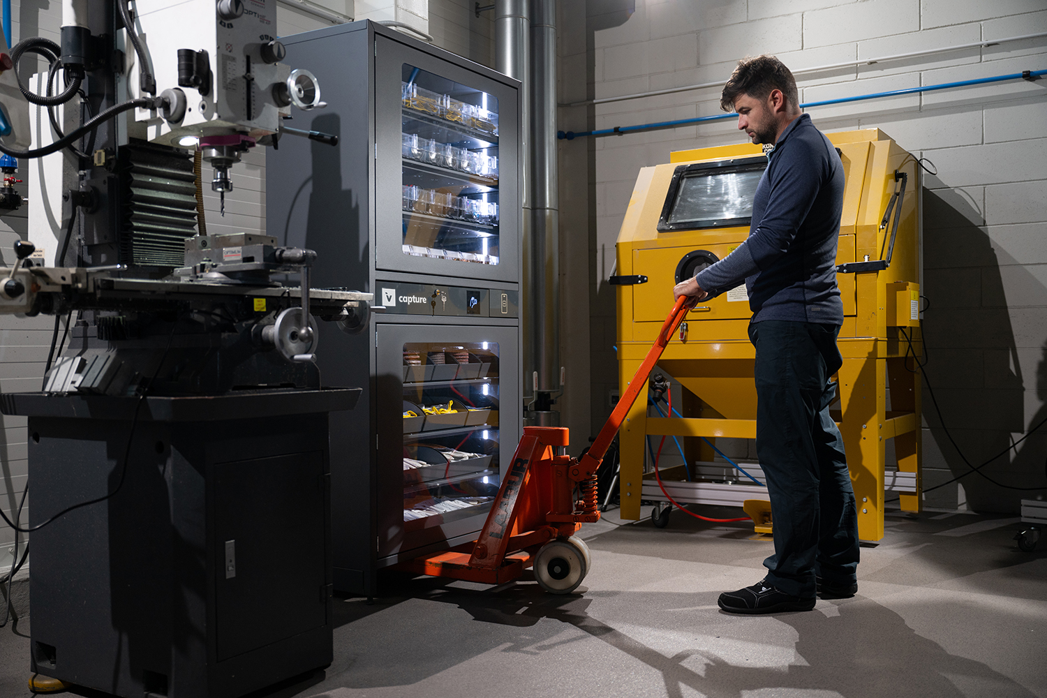 Man is using a forklift which is underneath an industrial vending machine, which interior LED lights are shining, in an industrial setting.