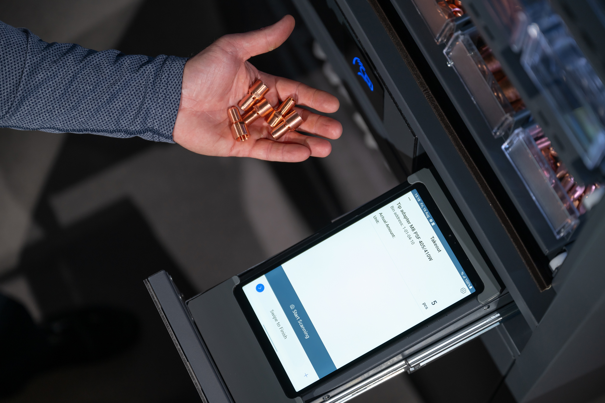 Close-up of a hand holding small copper parts above a tablet displaying an inventory management interface, with storage compartments visible nearby.