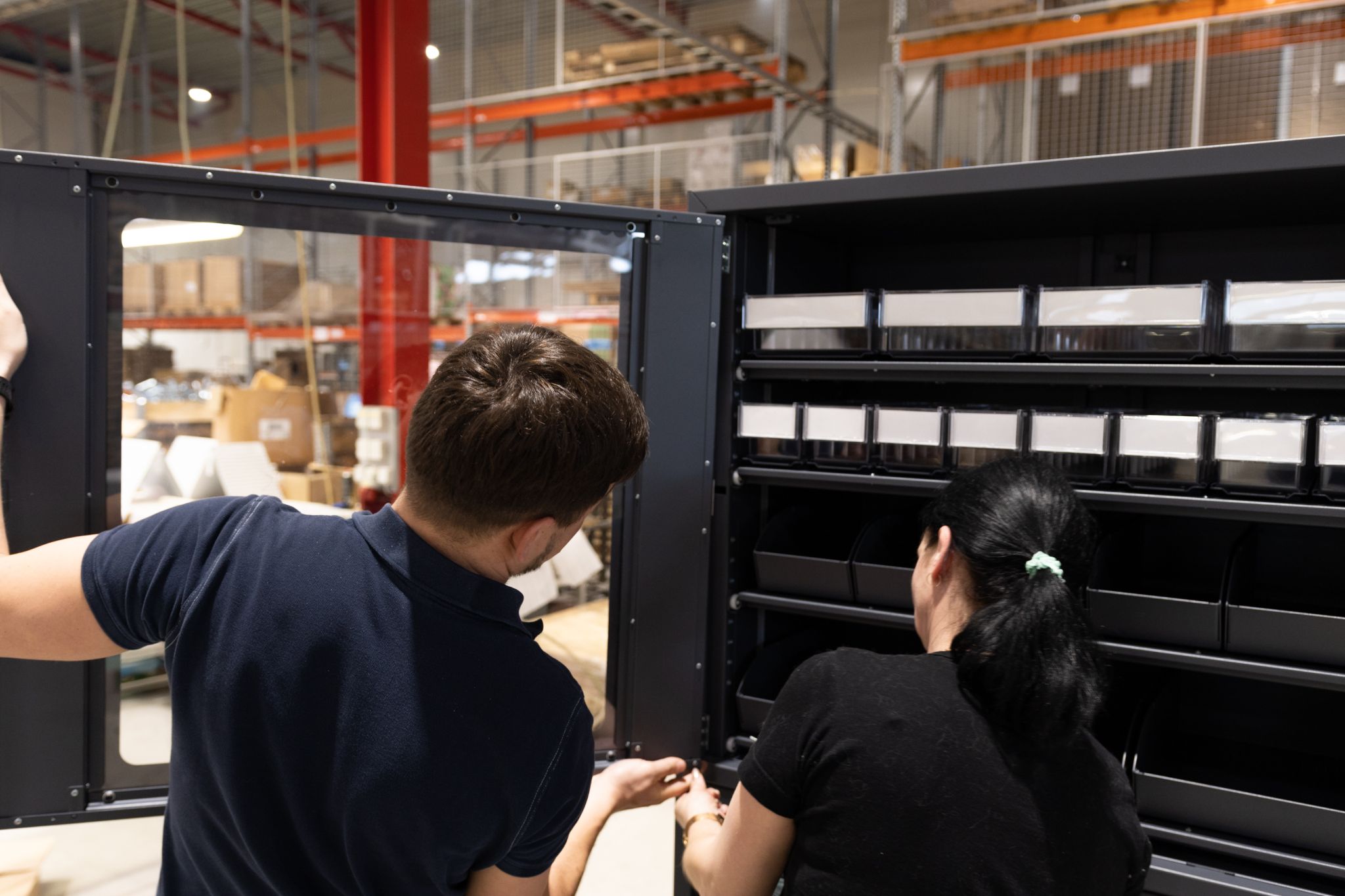 Two people inspecting a storage cabinet in an industrial warehouse, with shelves and equipment visible in the background.