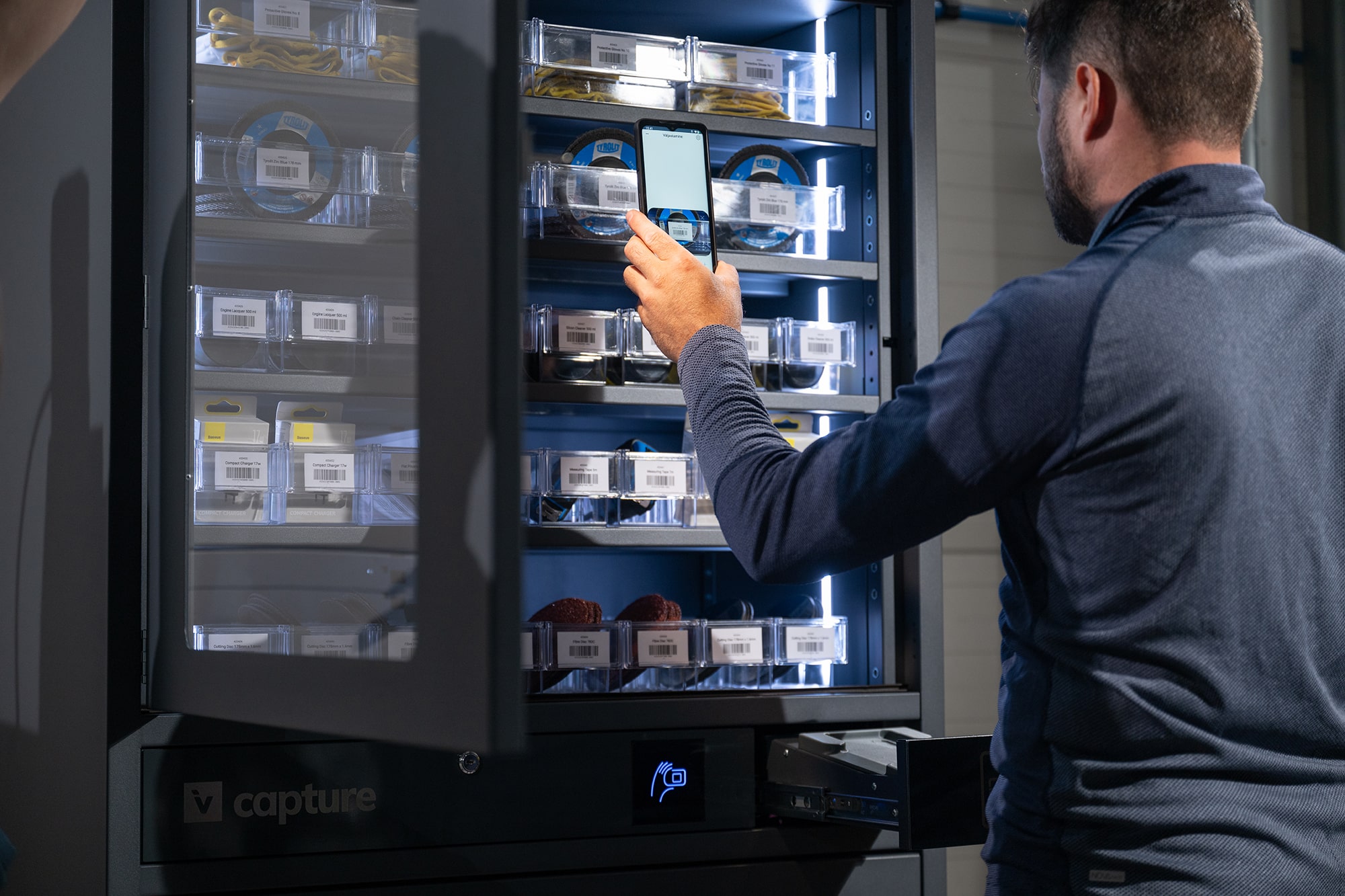 Man is standing in front of an industrial vending machine, the door of the vending machine is open and he is scanning the barcode on the shelf with a smartphone.
