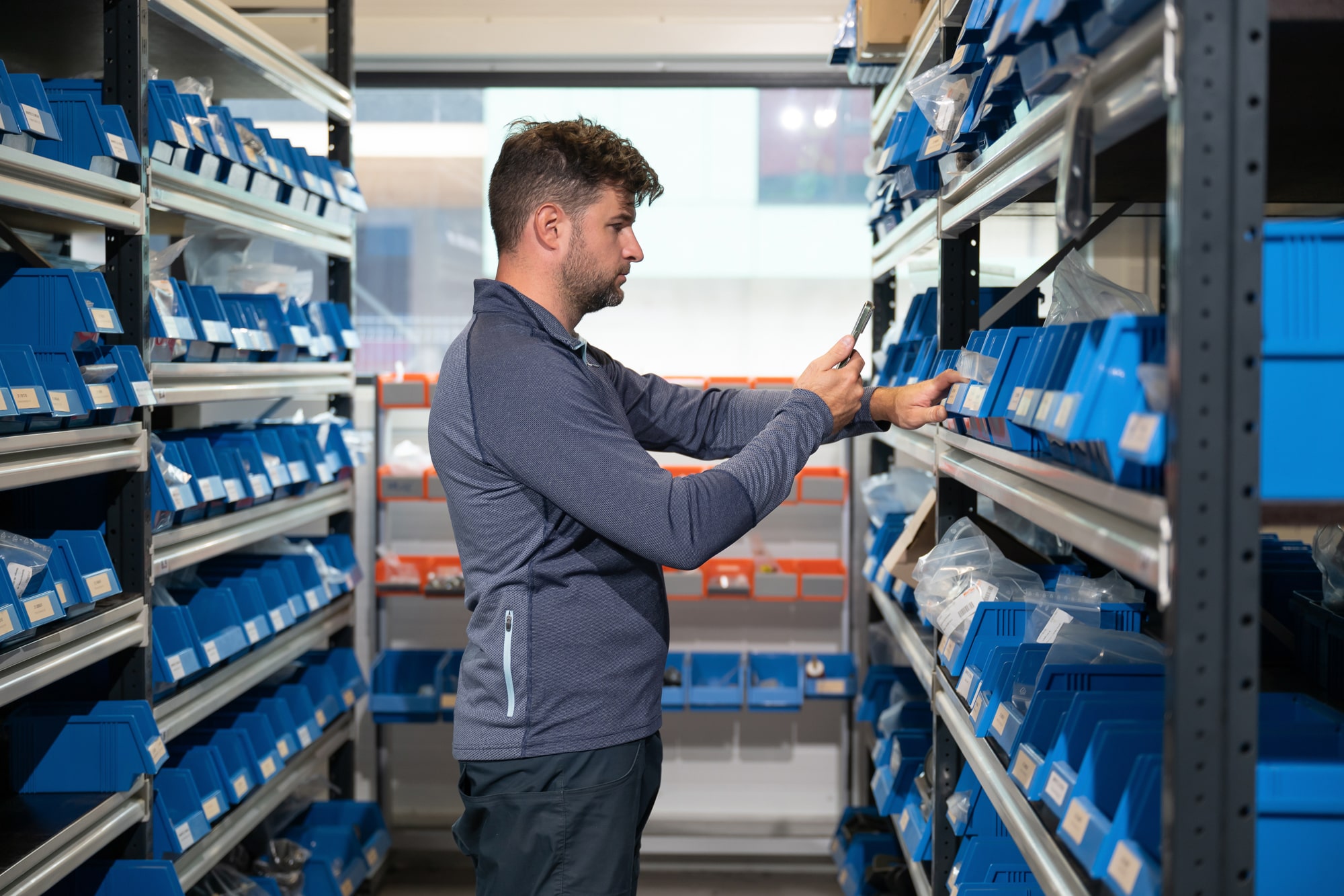 Man is standing between the shelves of an industrial environment, holding a smatphone and scanning an item from the shelf.