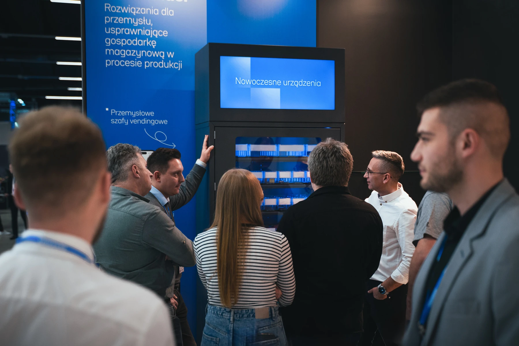 Group of people standing in front of a industrial vending machine and listen to a man who is giving a presentation.